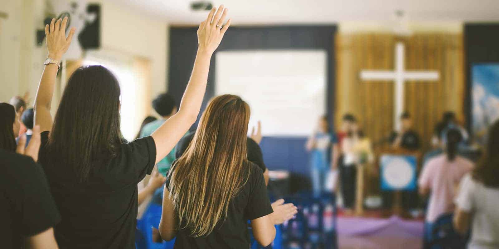 A woman lifting her hands and singing in worship with others in room with a white cross on the wall in the background.