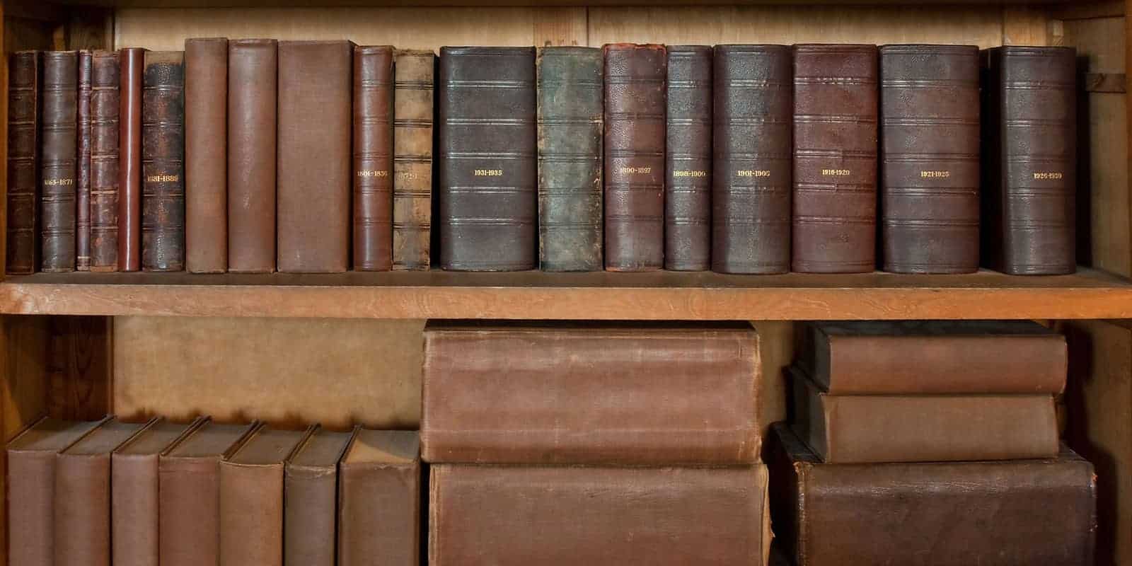 A picture of thick, old books stacked and piled on a bookshelf.