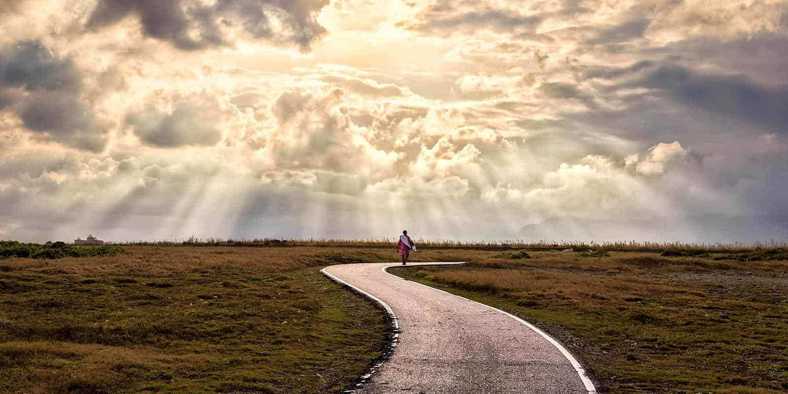 Beams of sunshine coming through the clouds shining down on a person walking on a winding path.