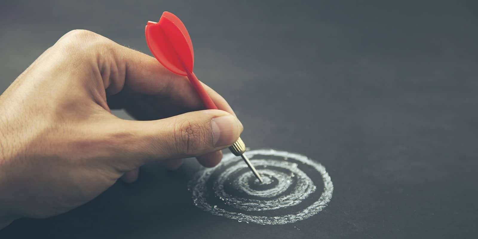 A person's hand holding a dart in the bullseye of a target.